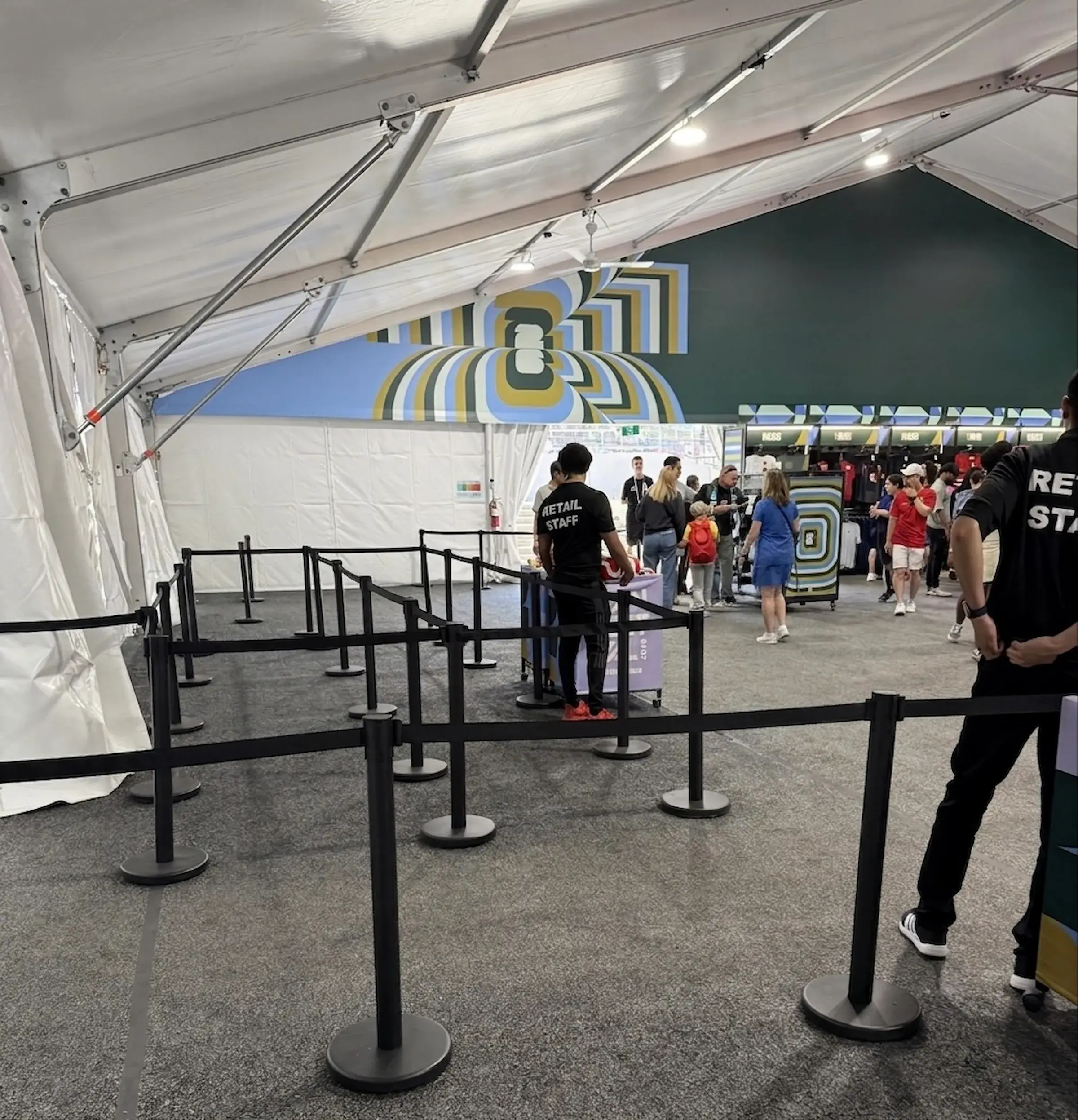 Black retractable-belt posts set out as queue lanes on grey carpet inside a white event marquee, with retail staff and a merchandise stand behind them